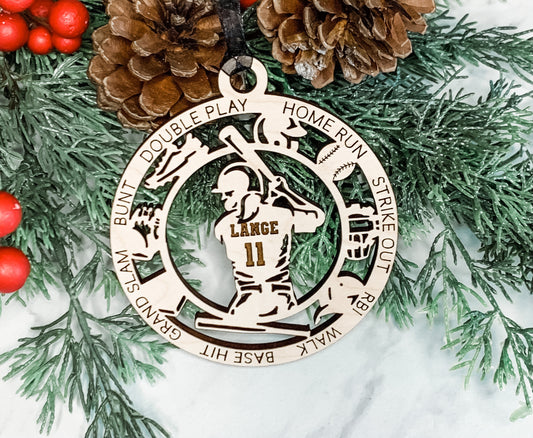 A personalized wooden ornament featuring a softball player, with the ability to have names and numbers engraved on it. It is displayed with a Christmas tree and pine cones in the background.